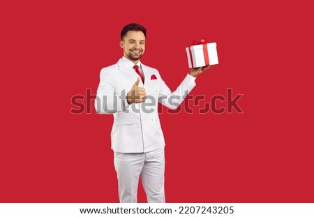 Smiling young man in suit isolated on red studio background hold present show thumbs up. Happy male in formal costume greeting congratulate with gift box recommend good service.
