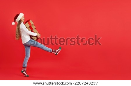 Full length profile side view happy excited child in cap and cute striped socks takes wide step while carrying stack of Christmas gift boxes on red copyspace banner background. Buying presents concept