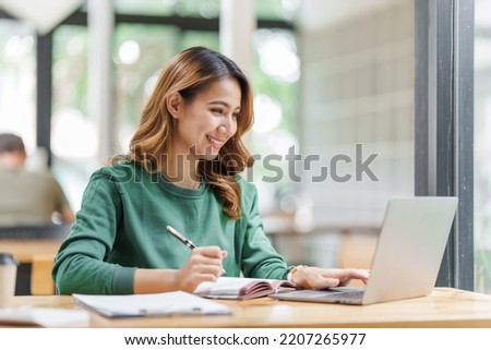 Happy asian business woman holding notebook paper and working with laptop in green sweater sitting at office desk.