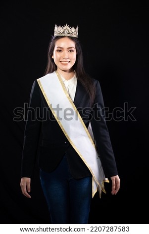 Half body portrait of Miss Pageant Beauty Contest in Asian smart suit blazer shirt dress with Silver Diamond Crown Sash, fashion make up smile wave hand, studio lighting black background isolated Royalty-Free Stock Photo #2207287583