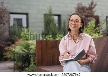 Portrait of confident smiling asian writer taking notes looking away sitting in park. Smart student wearing eyeglasses studying, reading book sitting in university campus. Education concept Royalty-Free Stock Photo #2207287967