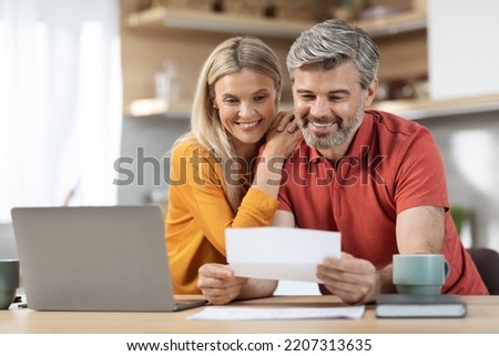 Happy middle aged spouses reading letter from bank, sitting at table in front of laptop, drinking coffee, checking correspondence together, kitchen interior, copy space. Mortgage, loan concept Royalty-Free Stock Photo #2207313635