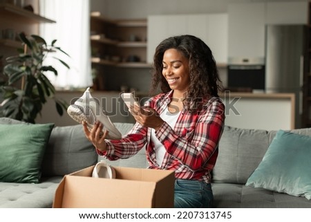 Glad young african american lady opens box, takes pictures of shoes on smartphone, enjoys online shopping in living room interior. Fashion blog, unboxing at home alone, app and sale for shopaholic