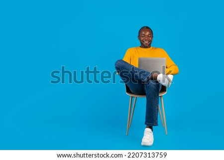 Handsome middle aged black man relaxing on chair with computer on his lap, looking at laptop screen and smiling, watching movie online, blue studio background, copy space