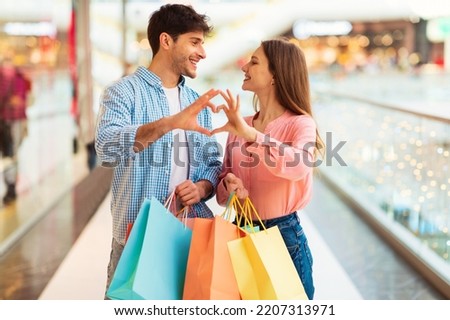 We Love Shopping. Spouses Making Fingers Heart Holding Colorful Shopper Bags Posing Smiling To Each Other Standing In Modern Mall Indoors. Valentine's Day Sales Offer Concept. Selective Focus Royalty-Free Stock Photo #2207313971