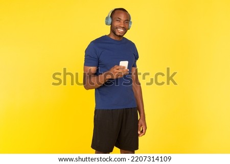 Cheerful African American Man Listening To Music Holding Cellphone Wearing Wireless Earphones Posing Over Yellow Studio Background, Wearing Sportswear. Great Audiobook Or Podcast Concept
