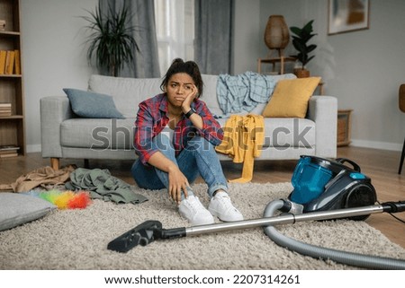 Unhappy tired young african american female sit on floor with vacuum cleaner, have problems with hygiene in living room interior. Domestic chores, mess, a lot of housework at home alone and disorder Royalty-Free Stock Photo #2207314261