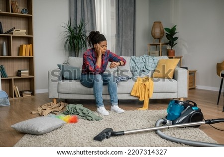 Need help with cleaning. Sad tired young black woman with vacuum cleaner looks at watch in living room interior. A lot of housework at home alone, chaos and disorder, stress, lifestyle and emotions Royalty-Free Stock Photo #2207314327