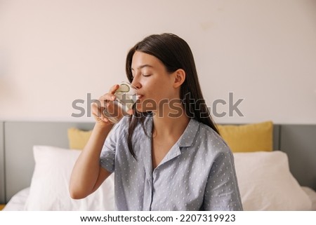 Pretty young caucasian woman drinking water with slice of cucumber sit on bed good morning. Brunette hair girl wears in grey pajamas. Concept healthy lifestyle