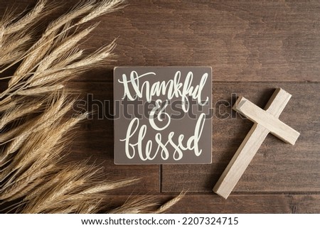 Words thankful and blessed with a border of wheat and a small wood cross on a dark wood background 