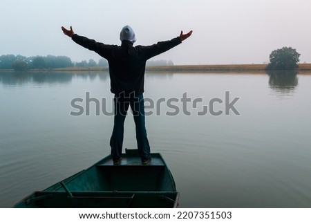 Young man in hoodie standing with hands up on fishing boat at foggy weather 