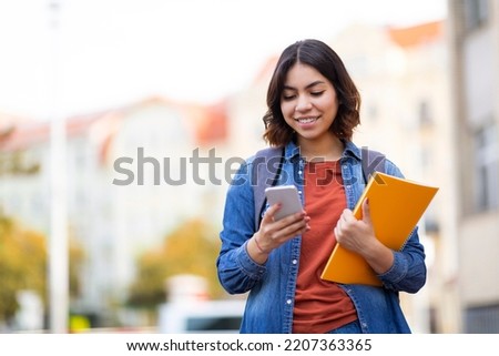 Smiling Young Middle Eastern Female Student Walking With Smartphone And Workbooks On City Street, Happy Millennial Arab Woman Messaging On Mobile Phone While Going Home After Classes, Copy Space Royalty-Free Stock Photo #2207363365