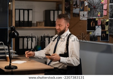 A professional detective works on a murder in the office at night. He sits at the table, holds a smartphone in his hands and reads the news online. copy space.
