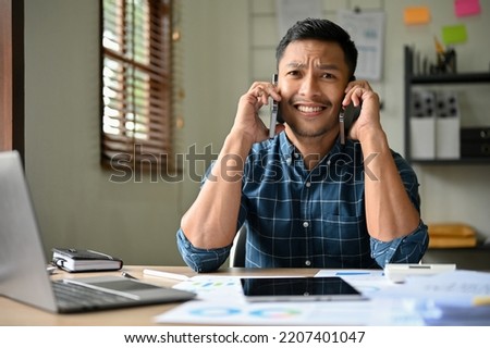 Confused and busy millennial Asian businessman or male office worker receives multiple phone calls at the same time while working in the office. Royalty-Free Stock Photo #2207401047