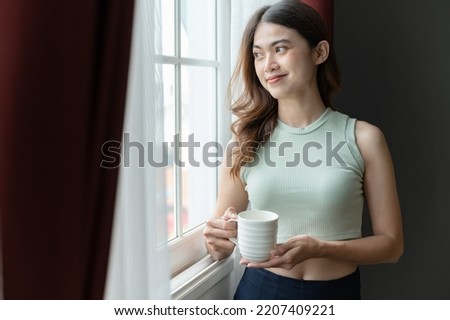 Happy Asia woman holding coffee in mug edge window in living room at home	