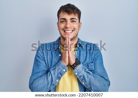Young hispanic man standing over blue background praying with hands together asking for forgiveness smiling confident. 