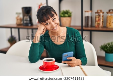 Young beautiful hispanic woman using smartphone drinking coffee sitting on table at home