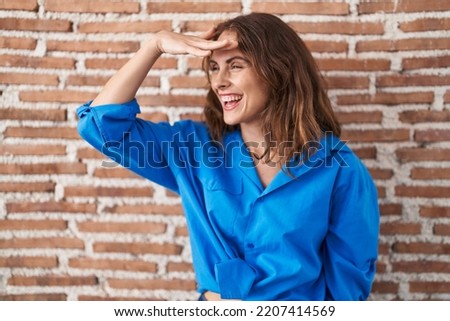 Beautiful brunette woman standing over bricks wall very happy and smiling looking far away with hand over head. searching concept. 