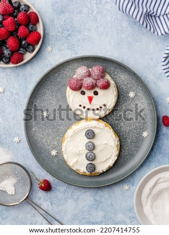 Pancakes snowman shaped with cream cheese frostind, fresh berries and sugar powder. Funny christmas breakfast or dessert for kids. Edible new year. Festive food concept. Top view. Blue background.