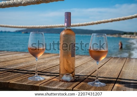 Glasses and bottle of cold rose wine from Provence served outdoor on wooden yacht pier with view on blue water and white sandy beach Plage de Pampelonne near Saint-Tropez, summer vacation in France
