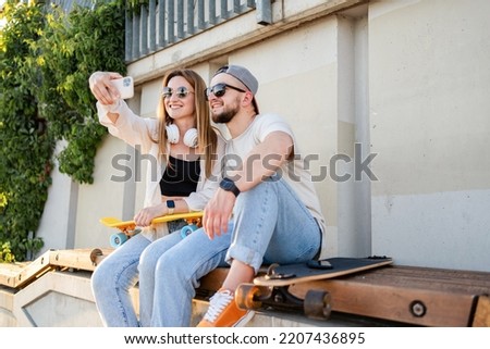 Pretty girl with headphones and young guy with skateboards taking photos on camera of cellphone together at street.