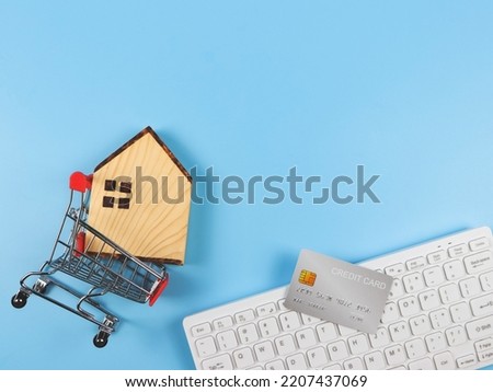 Top view or flat layout of wooden house model in shopping trolley, computer keyboard and credit card on blue background with copy space, home purchase concept.