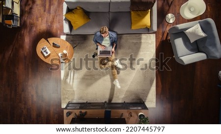 Wide Top View Apartment: Young Man Uses Laptop Sitting on a Carpet in Living Room. Creative Freelancer Working Remotely From Home. Stylish Entrepreneur doing E-Commerce Project, Online Business. Royalty-Free Stock Photo #2207445797