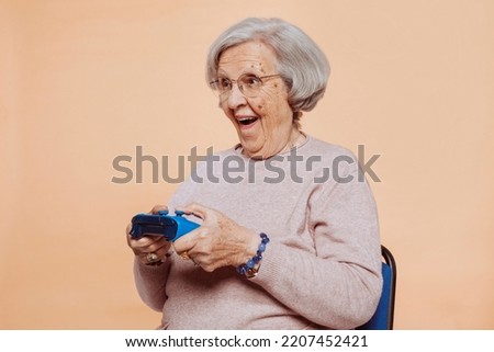 Close up portrait of excited elder grandmother with toothy smile playing video game at studio isolated over beige background. Player, console, gamer concept. Royalty-Free Stock Photo #2207452421