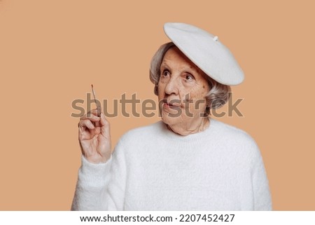 Close-up portrait of elder grandmother with grey hair, wears white sweater and beret holding brush isolated over beige background. Old female artist painter. 