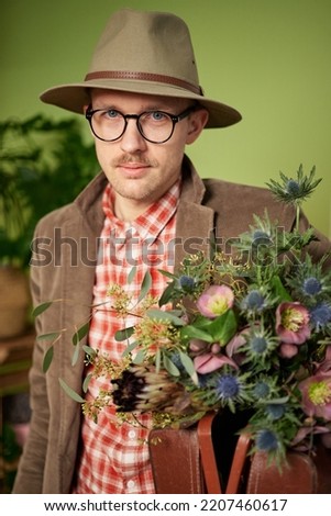 Cute old fashioned male person in eyeglasses, velvet jacket and hat staying with flower bouquet and briefcase in flower store. Looking at cameraHigh quality vertical image