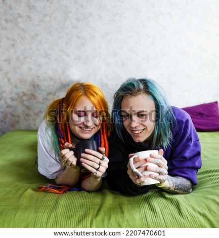 Portrait of a young tattooed punk couple of a girl and a guy with long dyed hair braided, lying on a large green bed. With stylish mugs in hand. Stylish modern youth