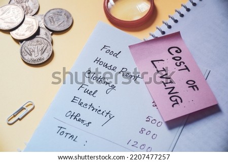 Cost of Living text with handwritten expenditure list and Indian currency coins on desk. Close-up, selective focus.