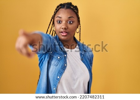African american woman with braids standing over yellow background pointing with finger surprised ahead, open mouth amazed expression, something on the front 