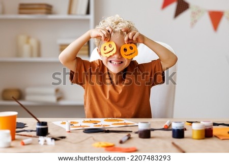 Portrait of cute kid  boy  making Halloween home decorations   while sitting at wooden table, child covers eyes with carved pumpkins and laughs merrily Royalty-Free Stock Photo #2207483293