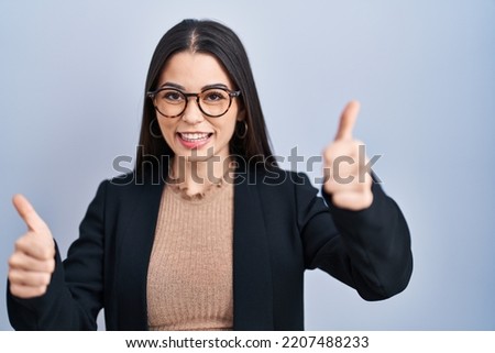 Young brunette woman standing over blue background approving doing positive gesture with hand, thumbs up smiling and happy for success. winner gesture. 