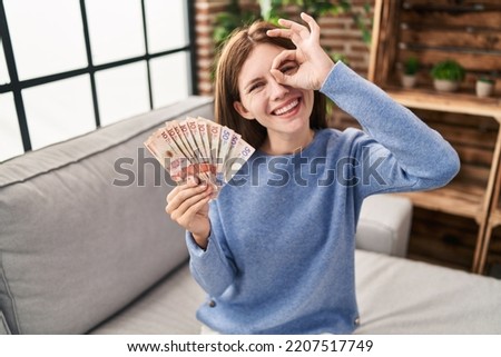 Young brunette woman holding colombian pesos smiling happy doing ok sign with hand on eye looking through fingers 