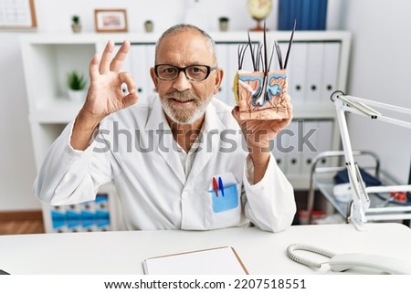 Mature doctor man holding model of human anatomical skin and hair doing ok sign with fingers, smiling friendly gesturing excellent symbol 