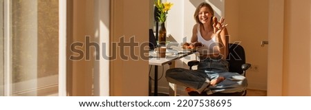 Smiling young woman holding croissant and looking at camera near gadgets and coffee at home, banner