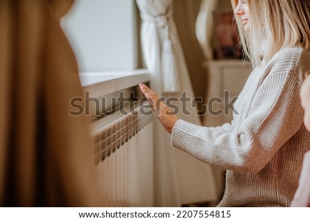 Young blond woman in long winter beige sweater is posing at home near the radiator. Winter season concept. Focus is at hands. Economy program Royalty-Free Stock Photo #2207554815