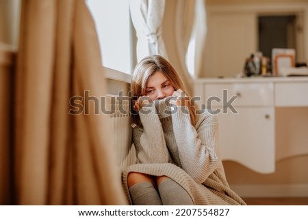 Young blond woman in long winter beige sweater is posing at home near the radiator. Winter season concept. Focus is at hands. Economy program Royalty-Free Stock Photo #2207554827