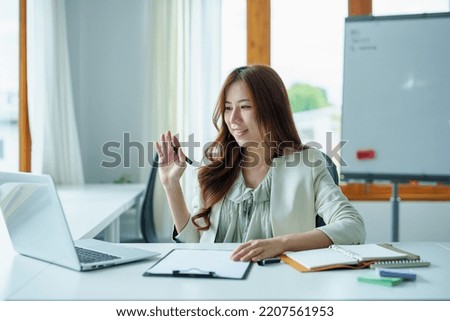 Portrait of an Asian bank employee using a laptop video conferencing with team members working on budget paperwork at their desk in their office.