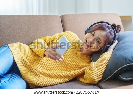 Portrait of a smiling Asian woman wearing a pair of headphones and using her phone and listening to music while sitting on the sofa.