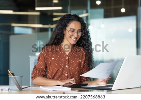 Young successful businesswoman behind paperwork, Hispanic woman working with documents and contracts inside modern office building, female worker using laptop, wearing glasses and curly hair smiling Royalty-Free Stock Photo #2207568863