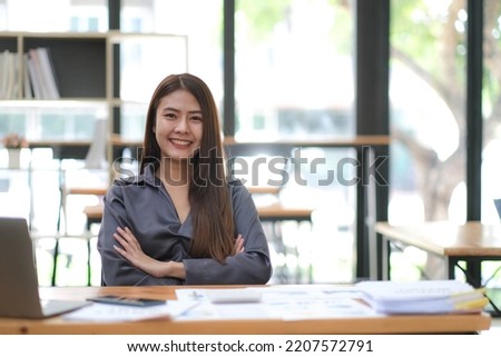 Confident young Asian business woman sitting with arms crossed smiling looking at camera in the office.
