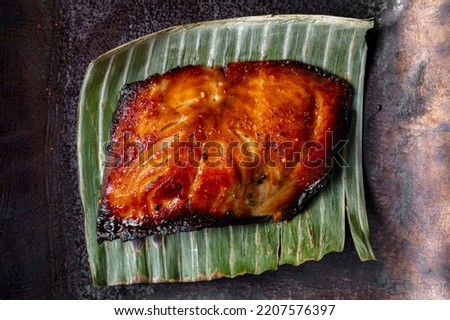 Grilled sea bass fillet in unagi sauce. The fillet lies on a palm leaf. Leaf and fish lie on a ceramic square plate. The plate stands on a gray stone background with bronze inlays.