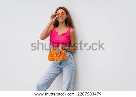Cute european woman in colorful summer outfit , pink crop top and blue jeans  posing over white background. Holding orange bag. Summer holidays  fashion concept. Royalty-Free Stock Photo #2207583479