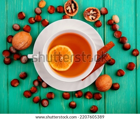 top view of cup of hot toddy with lemon inside and cinnamon on saucer with pattern of nuts and walnuts around on green background Royalty-Free Stock Photo #2207605389