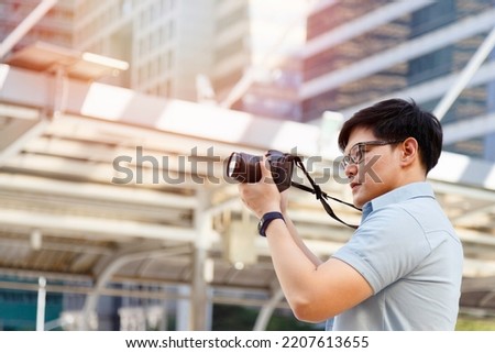 Young asian man photographer holding camera to taking picture of city view. Outdoor summer lifestyle portrait traveler having fun with shooting street photo in travel holiday vacation with sunlight.