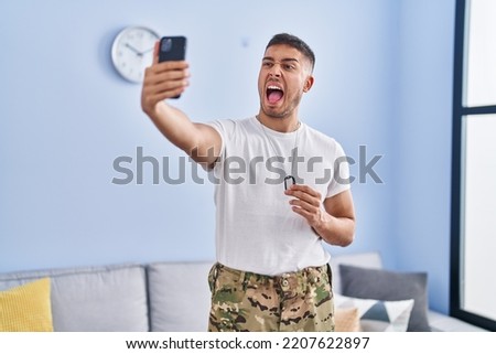 Young hispanic man wearing camouflage army uniform taking selfie at home angry and mad screaming frustrated and furious, shouting with anger. rage and aggressive concept. 