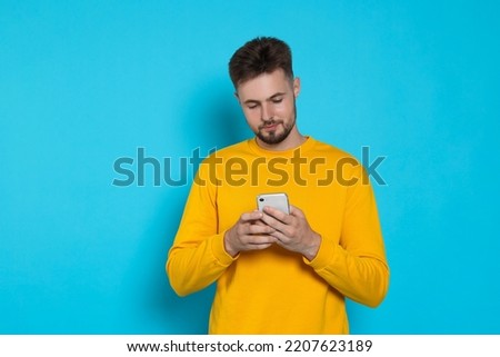 Handsome man in yellow sweatshirt with phone on light blue background